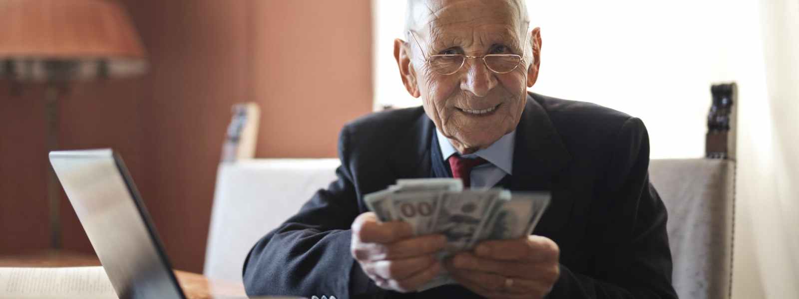 confident senior businessman holding money in hands while sitting at table near laptop