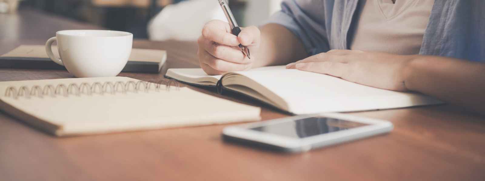 woman writing on a notebook beside teacup and tablet computer