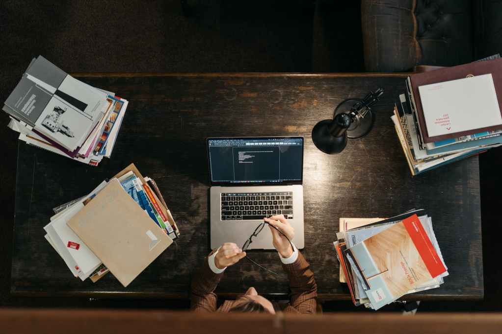 a student sitting behind a wooden desk with stacks of books and a laptop