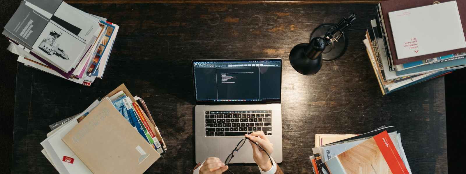 a student sitting behind a wooden desk with stacks of books and a laptop